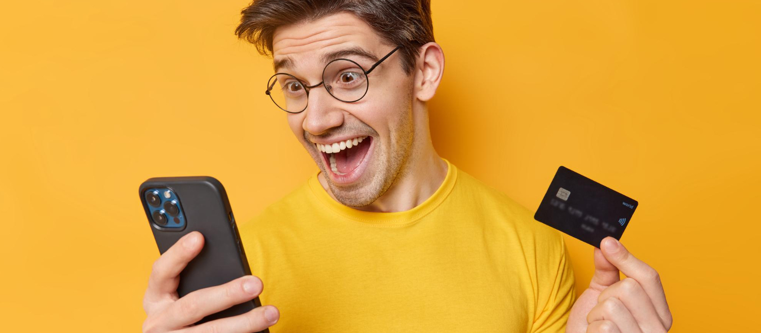 indoor-shot-positive-man-buys-something-online-holds-mobile-phone-plastic-card-glad-get-lump-sum-money-his-bank-account-wears-round-spectacles-casual-t-shirt-isolated-yellow-wall
